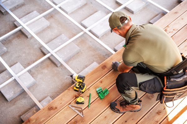 A construction worker focuses on laying down wooden planks to create a sturdy floor in a home. Tools such as a drill, tape measure, and square rest on the wooden surface. The worker is dressed in functional attire, showcasing dedication to precise craftsmanship.