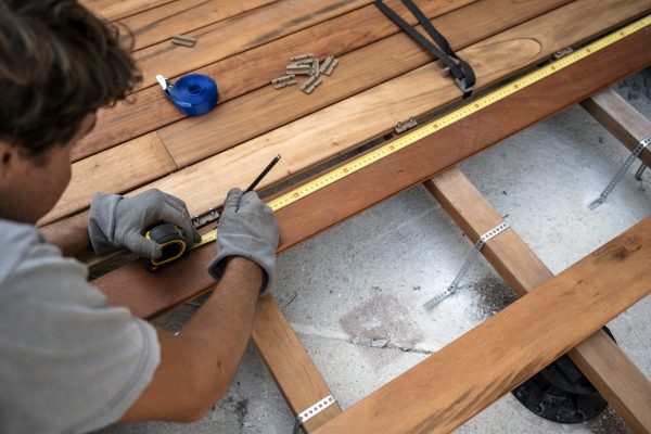 A craftsman measures and marks a wooden deck plank with a tape measure and pencil, ensuring accuracy during construction.